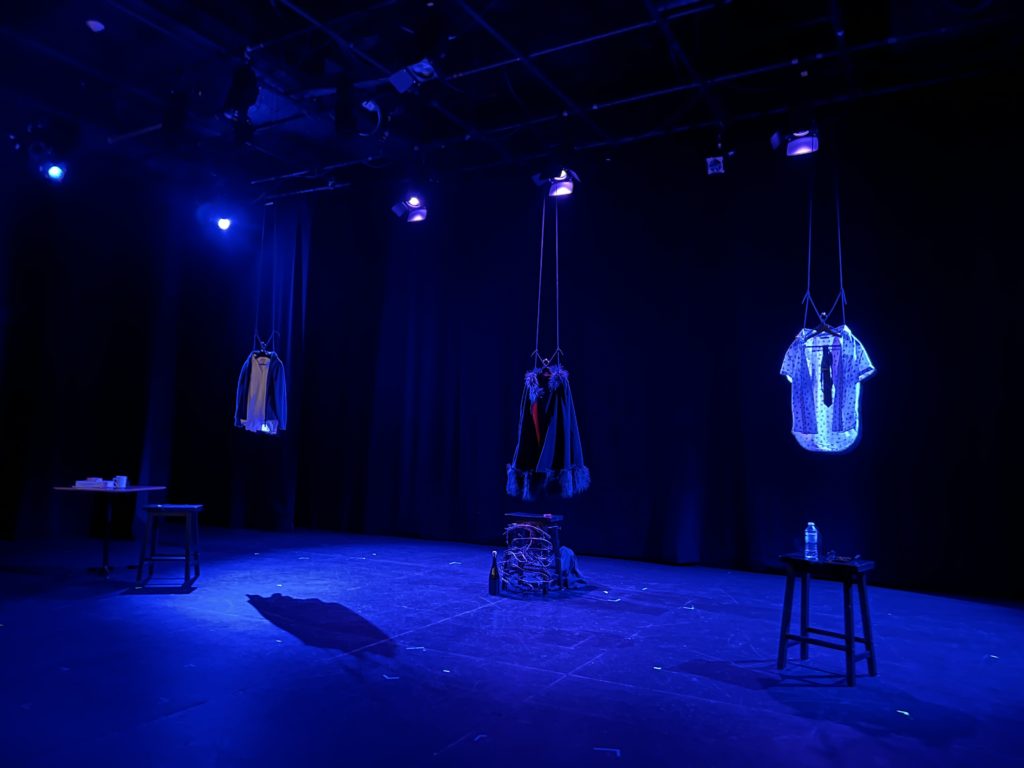 Three sett of clothing hang above a dark stage, bathed in blue light and framed by small white lights