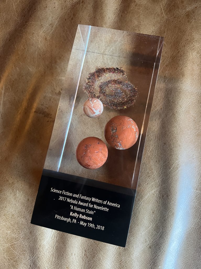 Rectangular clear lucite with three round, orange stones representing planets, with a swirl of orange and brown sand above. Black lower portion is inscribed: 

Science Fiction and Fantasy Writers of America
2017 Nebula Award for Novelette
"A Human Stain" 
Kelly Robson
Pittsburgh, PA - May 19th, 2018