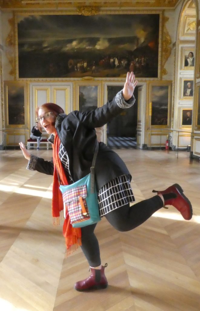 In a formal Versailles reception room, with many paintings on the surrounding walls, Kelly (a red haired woman in glasses, wearing a black and white dress, black leggings, black jacket, and red Blundstone boots) strikes a goofy, one legged pose.