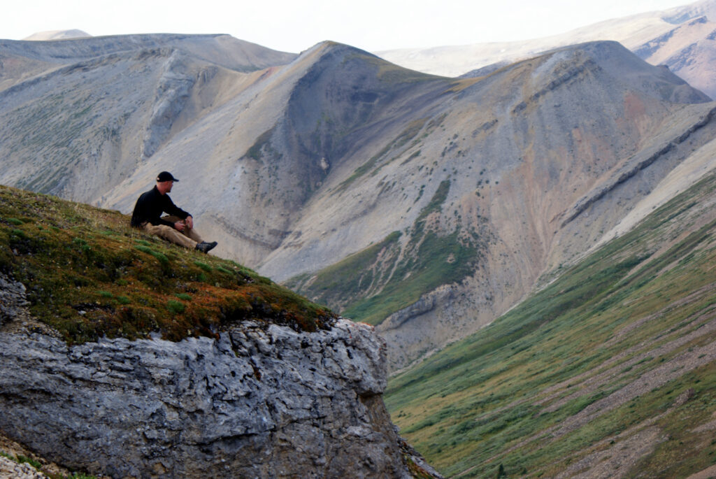 A cliff with a man sitting on it, looking away from the camera. In the background is a high mountain ridge, very eroded, in colors of peach, blue, and green