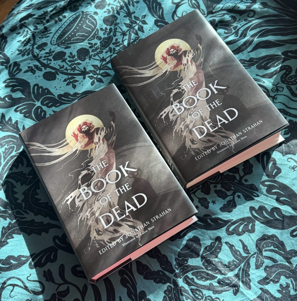 Two copies of a hardcover book like on a teal and black cloth. The covers say THE BOOK OF THE DEAD edited by Jonathan Strahan and illustrated by Galen Dara. The cover has an image of a spooky ghost-like figure with trailing hair and smoke in front of a full moon. It has a skull face and roses in its hair. Perhaps it is a ghost bride. The silhouette of a headstone is in the background.